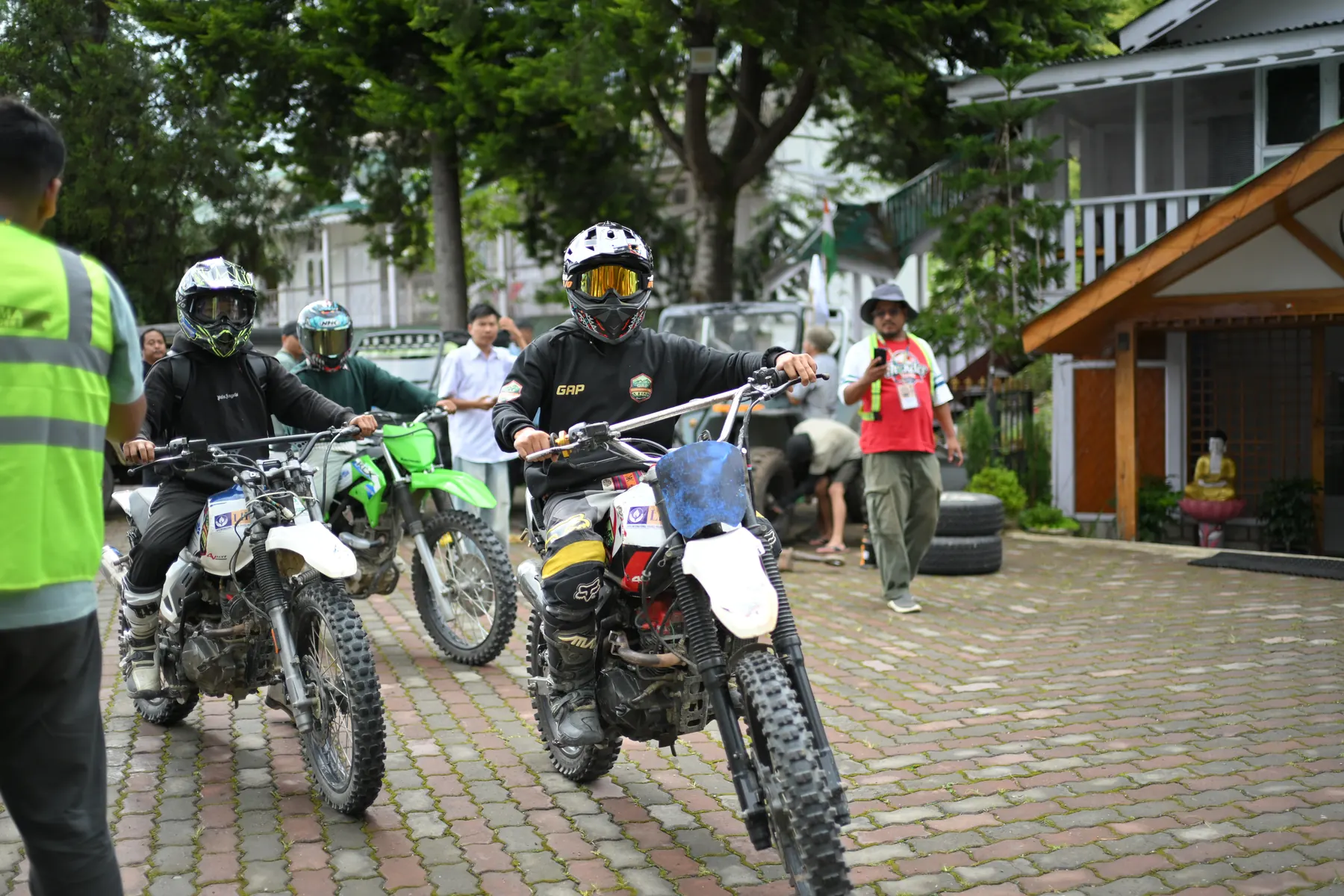 Riders on dirt bikes lined up on a paved courtyard beside wooden cottages, with a marshal in a high-visibility vest.