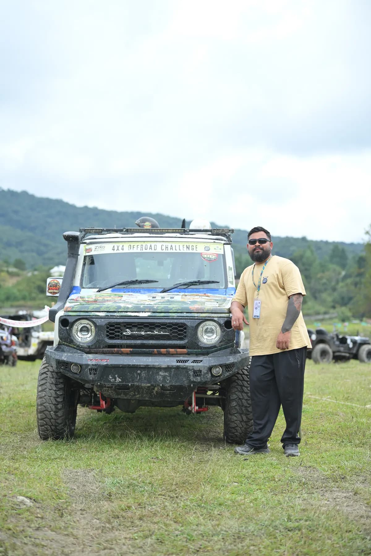 A man standing beside a mud-spattered off-road SUV on a grass field, hills behind.
