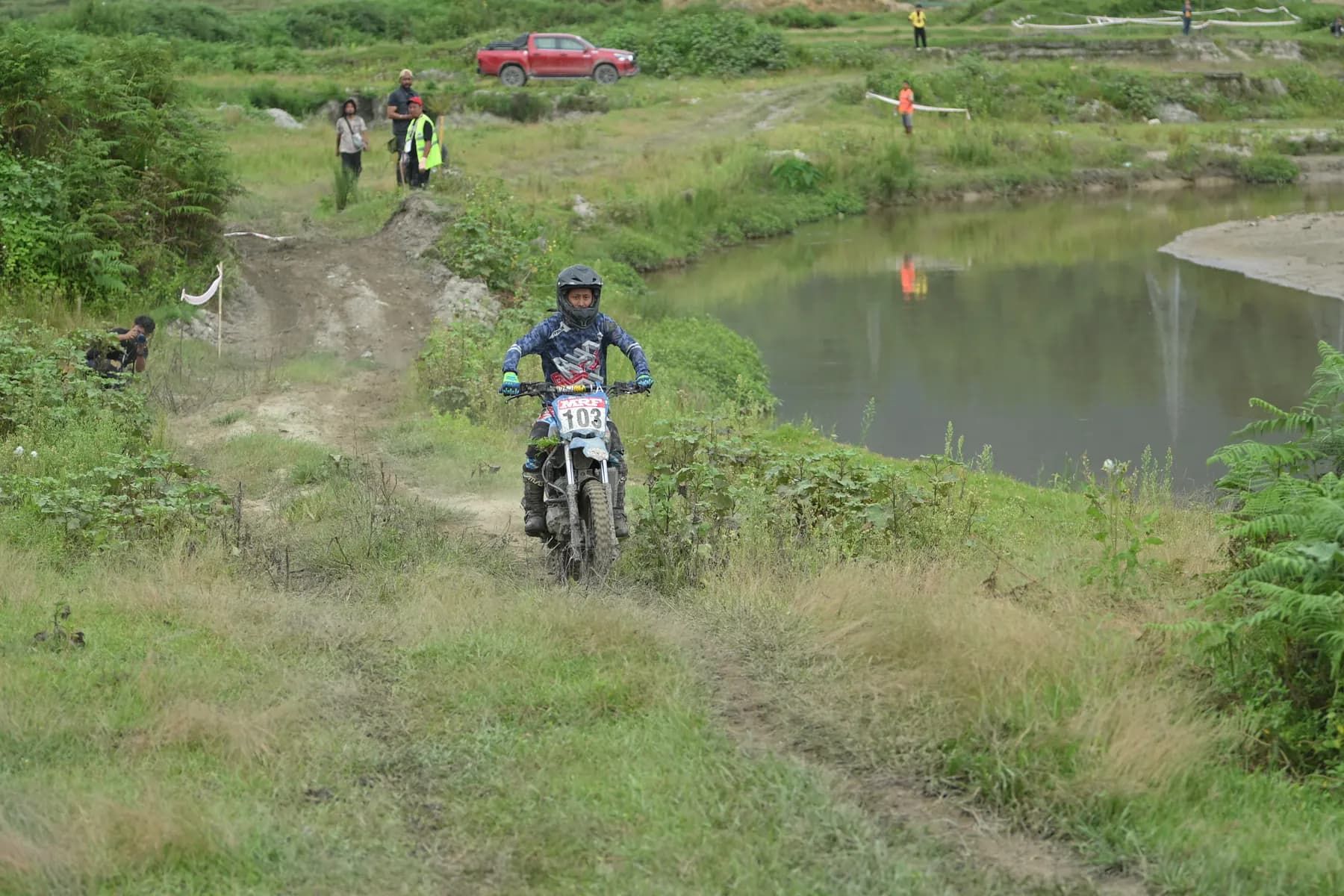 A rider on a dirt bike on a grassy trail beside a pond, marshals in the distance.