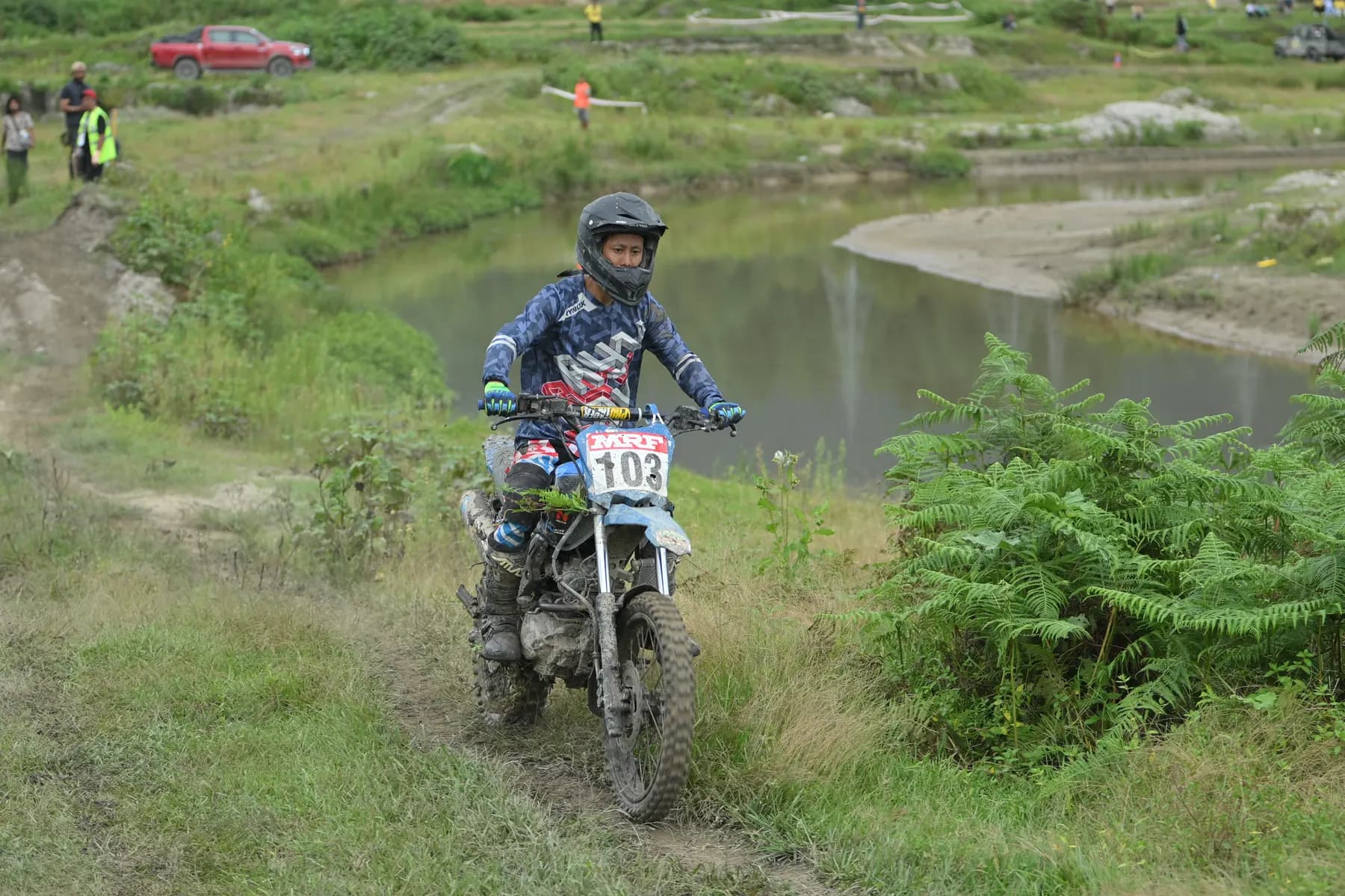 Rider number 102 on a dirt bike on a grass trail beside a pond.