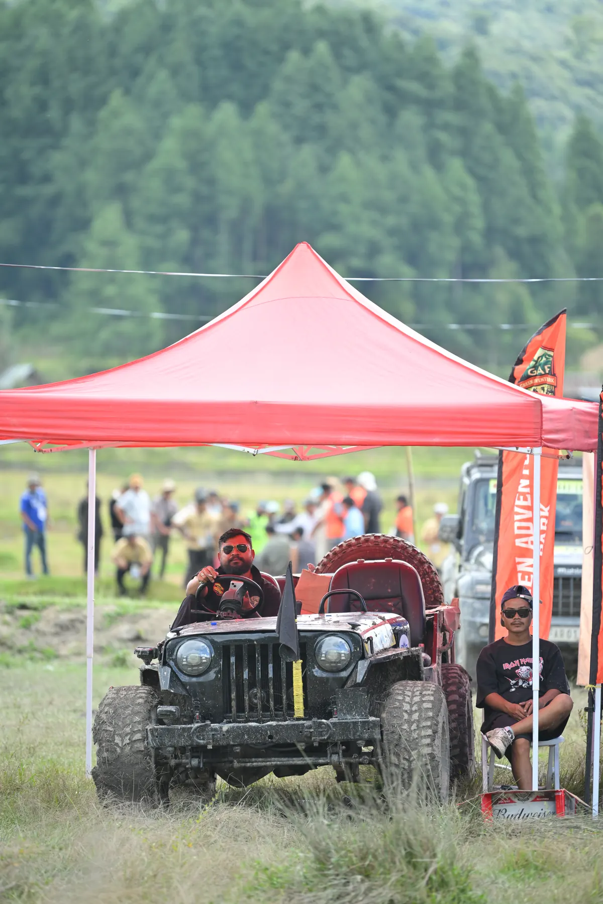 An off-road jeep under a red canopy, with spectators gathered behind.