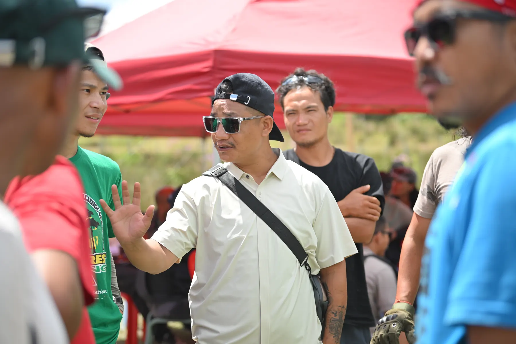 A man in a cream shirt speaking with a group under red canopies.