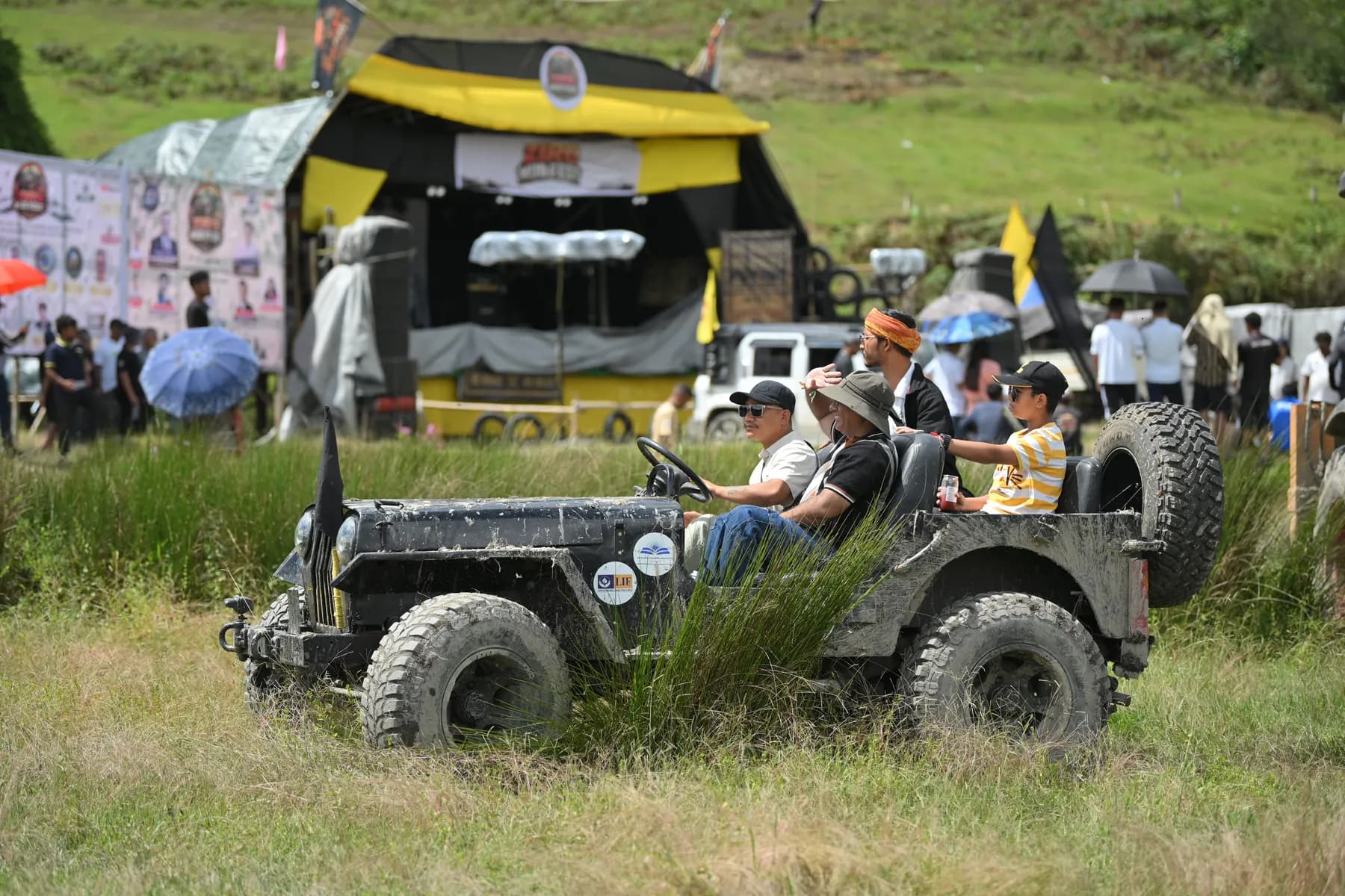 An open jeep with flags crossing tall grass in front of the stage area.